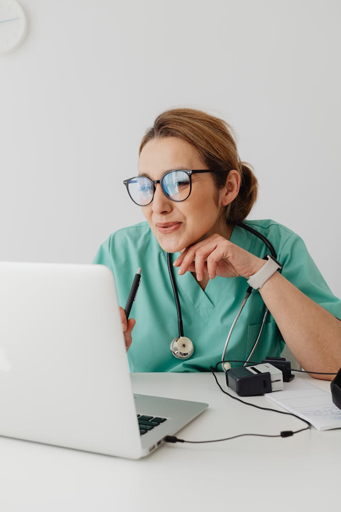 hero-gallery-04 A healthcare professional wearing scrubs and eyeglasses using a laptop with a stethoscope around the neck.